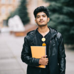 handsome-young-indian-student-man-holding-notebooks-while-standing-street